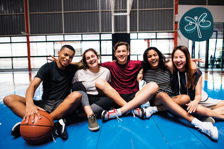 A group of smiling young adults sitting together in a gym after a game of basket ball